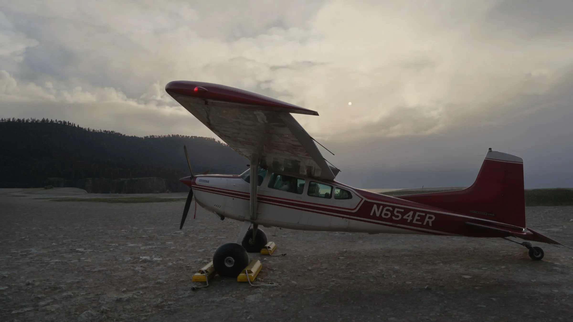 Red and white Cessna 185 Skywagon parked on a gravel strip at dusk beneath a heavy overcast, forested ridge and rising moon behind, rendered with REX Atmos CORE in Microsoft Flight Simulator 2024